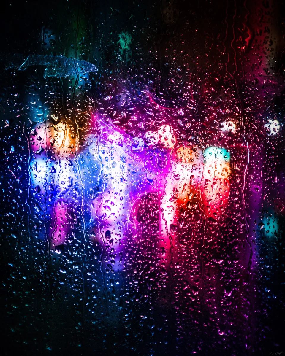 Raindrops on glass in front of blurred pink, red and blue city lights at night