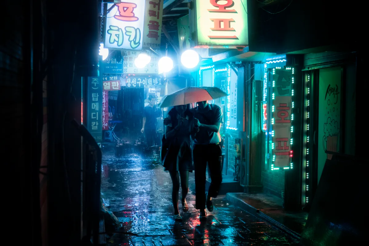 Two people sharing an umbrella under teal and red neon signs on a rainy Seoul street