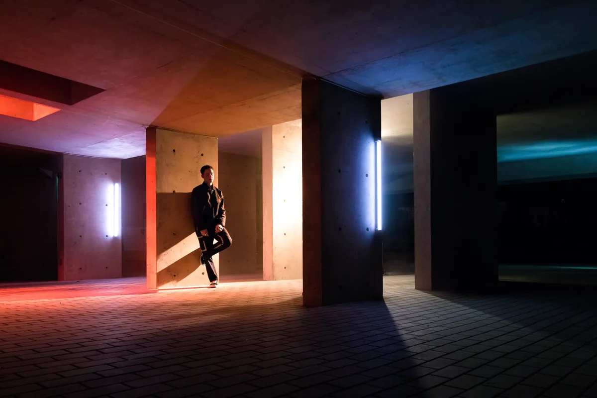 A figure leaning in a concrete underpass lit by orange light and a cold blue glow in Toronto