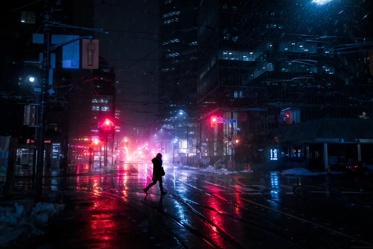 Between, a figure crossing a wet street under pink and red light at night in Toronto's Financial District